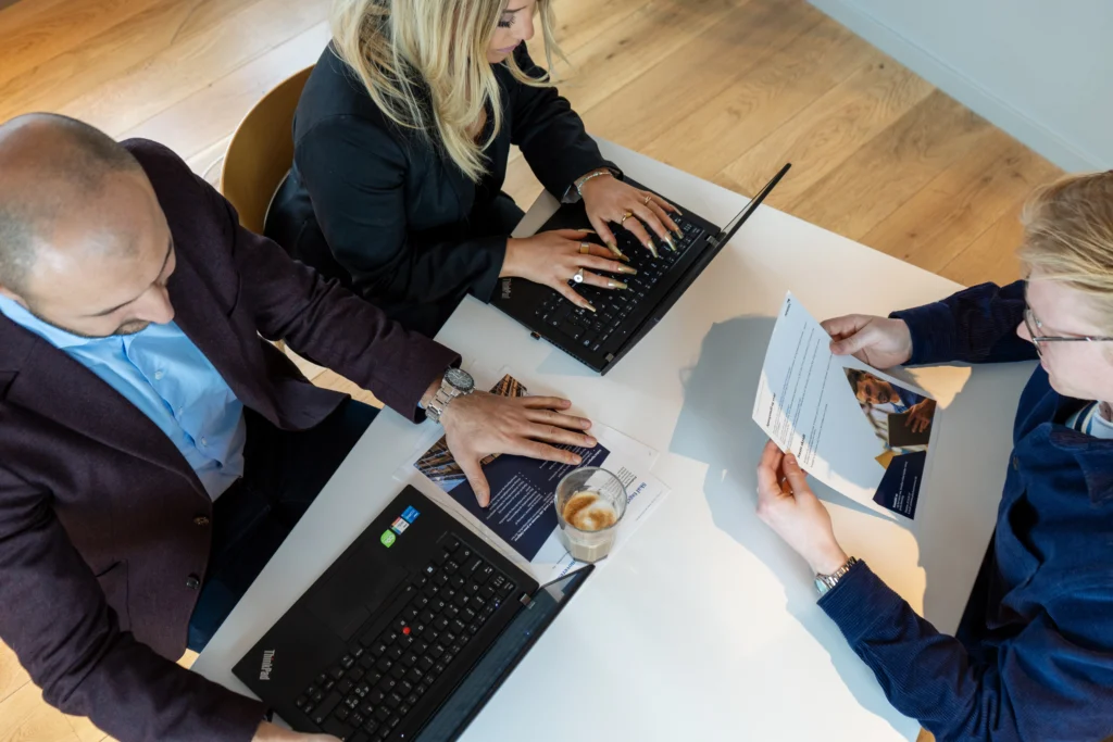 Tre personer ved mødebord med laptops og papirer, der planlægger ejendomsadministration og foreningsadministration for en ejerforening